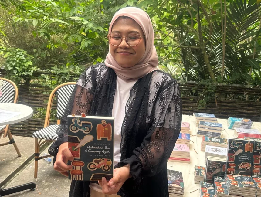 Woman in a pink hijab and glasses proudly holding a book outdoors, with a table of books in the background.