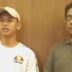 Two men stand indoors against a wooden wall; left is in a white cap and white shirt with a logo, right wears glasses and a green polo.