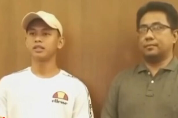 Two men stand indoors against a wooden wall; left is in a white cap and white shirt with a logo, right wears glasses and a green polo.