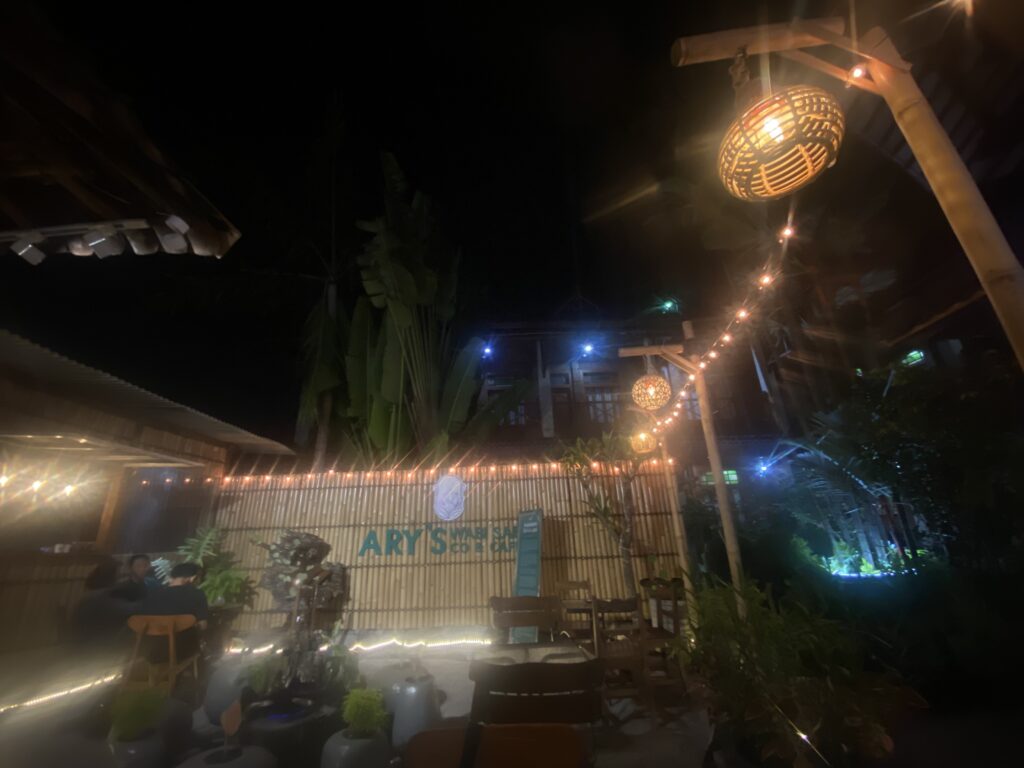 Outdoor night scene with warm hanging lanterns and string lights over a courtyard patio with tables and chairs.