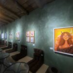 Gallery interior with green textured wall, wooden ceiling beams, and a large orange portrait on the right.