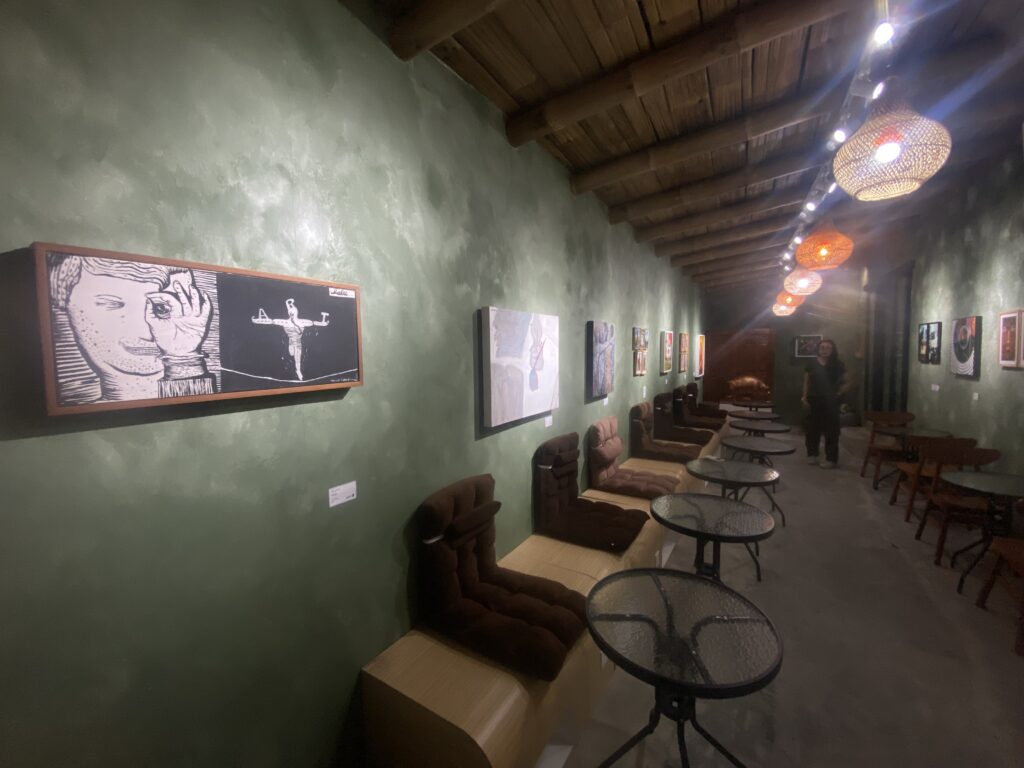 Green textured gallery wall with framed artwork, wooden ceiling beams, and hanging wicker pendant lights along a row of round glass tables and chairs on the floor level in an art space.