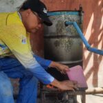 Man wearing a yellow shirt and black cap crouches beside a weathered metal drum with a blue pipe, pouring from a pink pitcher into a small stove underneath.