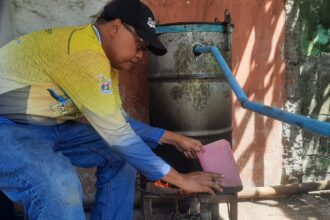 Man wearing a yellow shirt and black cap crouches beside a weathered metal drum with a blue pipe, pouring from a pink pitcher into a small stove underneath.