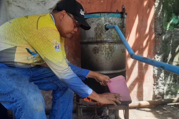 Man wearing a yellow shirt and black cap crouches beside a weathered metal drum with a blue pipe, pouring from a pink pitcher into a small stove underneath.