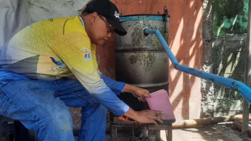 Man wearing a yellow shirt and black cap crouches beside a weathered metal drum with a blue pipe, pouring from a pink pitcher into a small stove underneath.