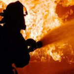 Firefighter in full gear silhouetted while spraying water at a large blaze in orange flames behind him