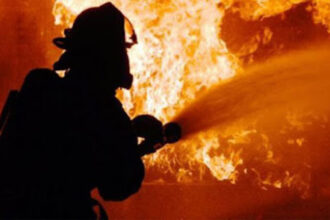 Firefighter in full gear silhouetted while spraying water at a large blaze in orange flames behind him