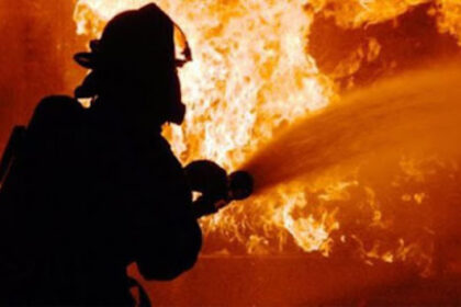 Firefighter in full gear silhouetted while spraying water at a large blaze in orange flames behind him