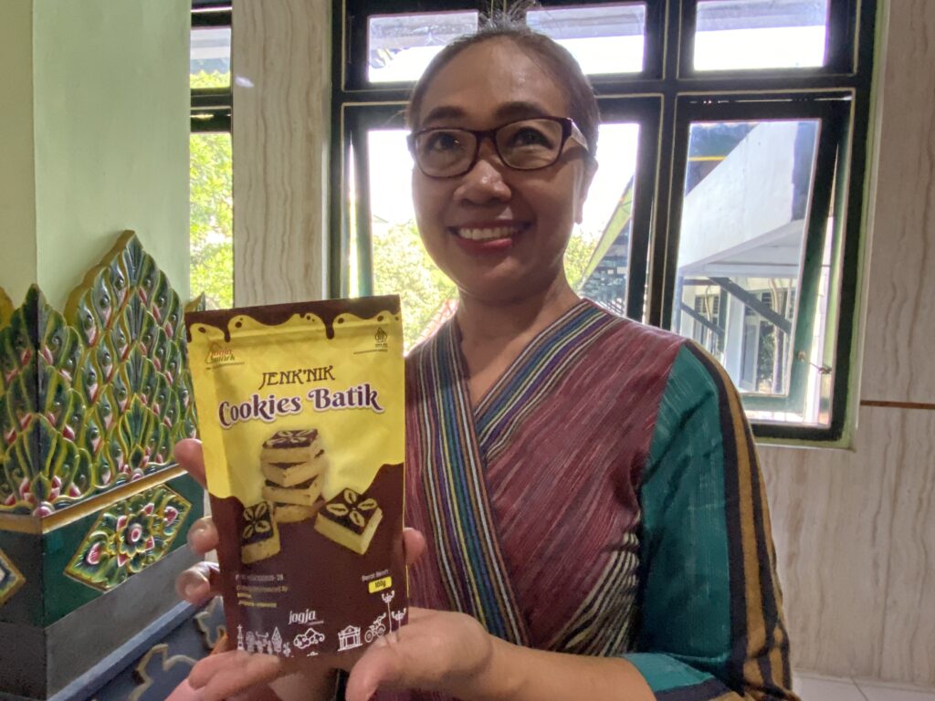 Smiling person with glasses holding a brown Batik cookies box indoors, colorful background behind them.
