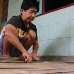 Man crouching on a wooden deck, smoothing or cleaning bamboo planks beside a tiled wall.