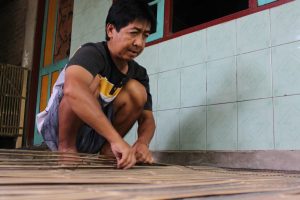 Man crouching on a wooden deck, smoothing or cleaning bamboo planks beside a tiled wall.