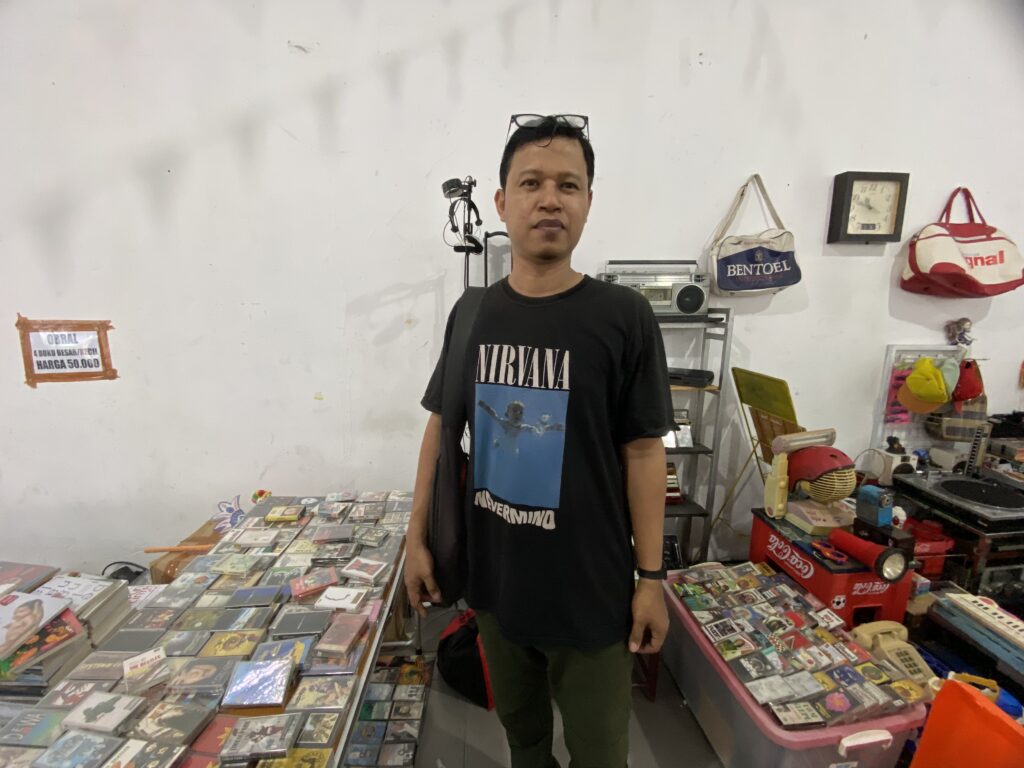 Man wearing a Nirvana T-shirt stands in a cluttered shop with shelves of CDs and vintage items in the background.