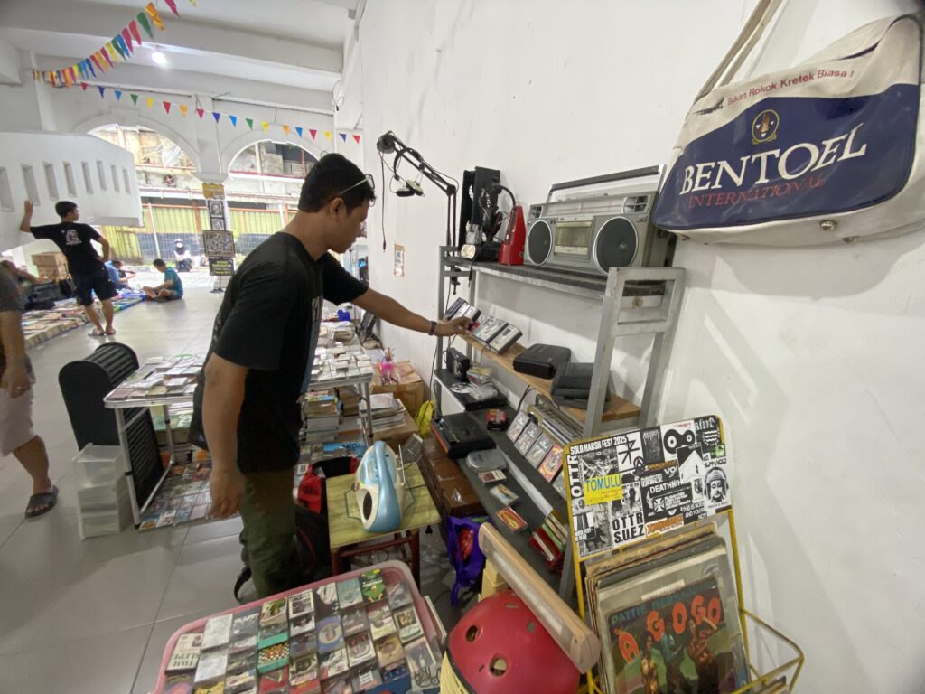 Inside a bright market stall with tables and racks of magazines and compact discs; a man in a black shirt browses the items while others browse in the background, colorful pennant banners overhead.