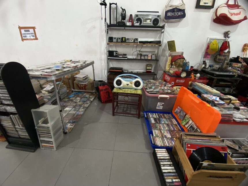 Cluttered room with shelves holding old electronics, cassette tapes, and CDs scattered on tables and shelves.