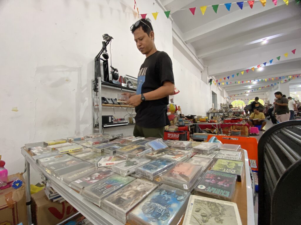 Man wearing a black T-shirt browses vinyl records at a booth, with shelves of records and colorful pennant banners above.