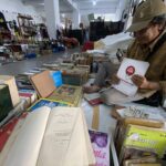 Man in a brown plaid shirt and cap sits among stacks of books and magazines in a busy indoor flea market or secondhand bookstore, handling a brochure.