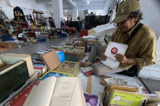 Man in a brown plaid shirt and cap sits among stacks of books and magazines in a busy indoor flea market or secondhand bookstore, handling a brochure.