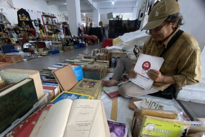 Man in a brown plaid shirt and cap sits among stacks of books and magazines in a busy indoor flea market or secondhand bookstore, handling a brochure.