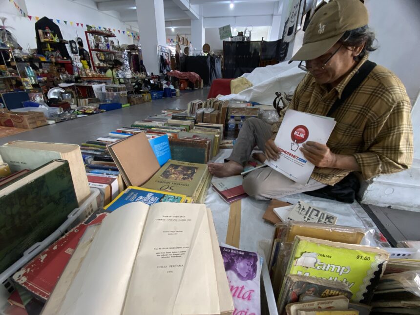 Man in a brown plaid shirt and cap sits among stacks of books and magazines in a busy indoor flea market or secondhand bookstore, handling a brochure.