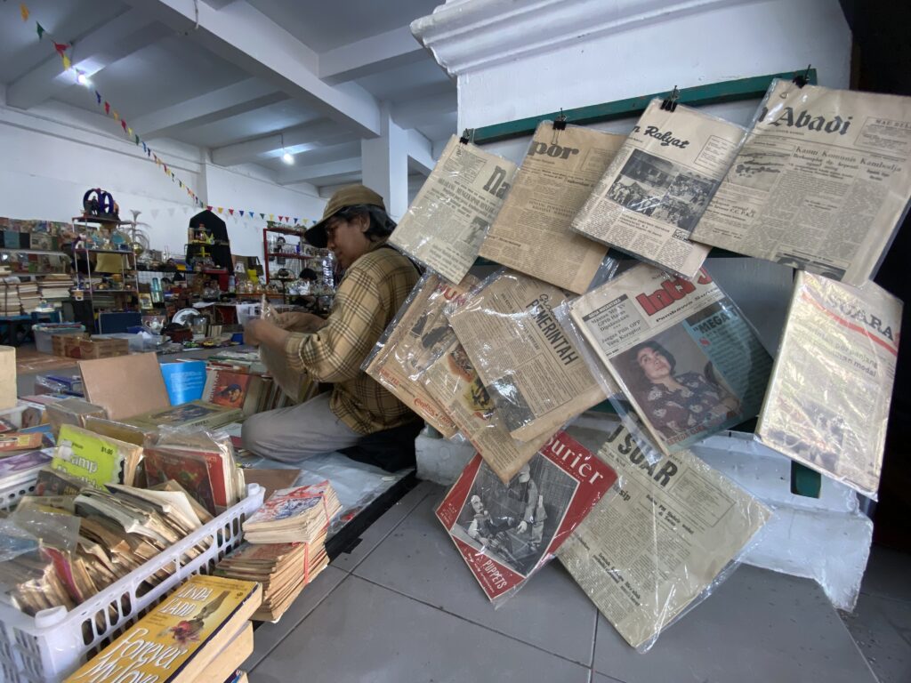 Man in a plaid shirt and cap sorting through newspapers in a cluttered bookstore.