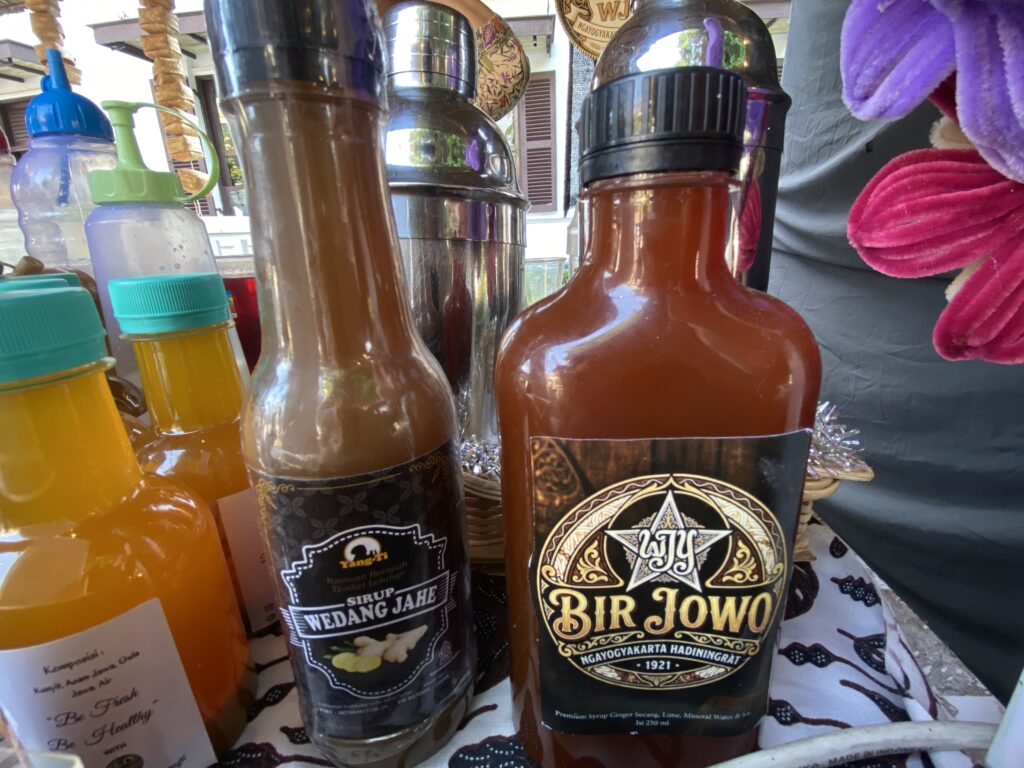 Close-up of several glass bottles filled with orange to dark brown liquids at a market stall, with decorative labels and a purple towel in the background.