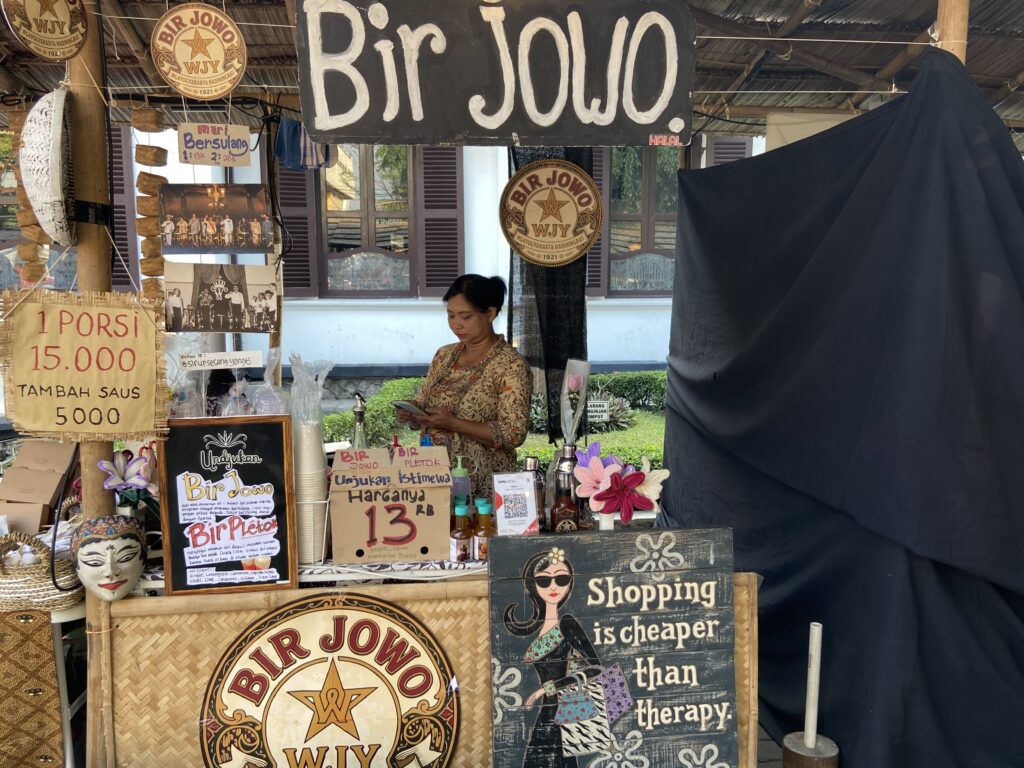 Market stall selling Bir Jowo items with signs, photos, and a woman behind the counter.