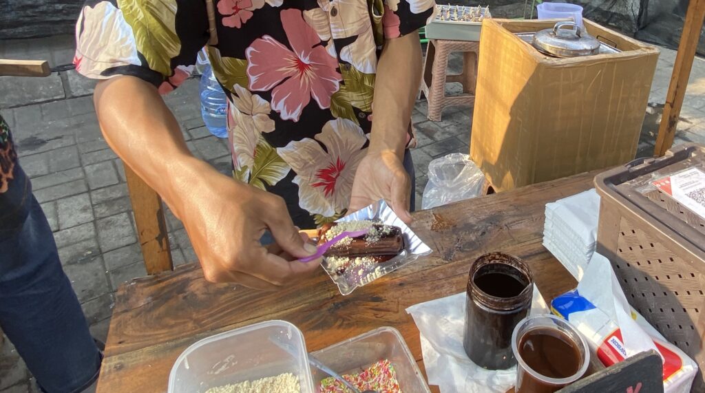 Person decorating a chocolate pastry with sprinkles on a foil-lined tray at an outdoor stall.