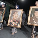 Nighttime street procession: three people carry large framed portraits adorned with garlands, walking barefoot or in traditional clothing.
