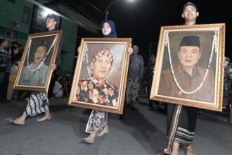 Nighttime street procession: three people carry large framed portraits adorned with garlands, walking barefoot or in traditional clothing.