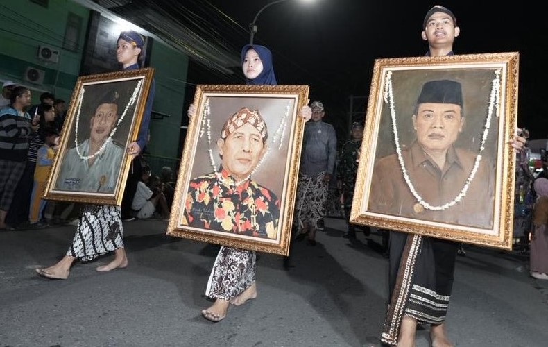 IMG_9082 - Mabur.co Nighttime street procession: three people carry large framed portraits adorned with garlands, walking barefoot or in traditional clothing.