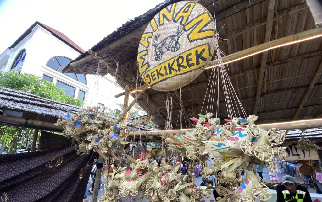 Round woven sign with yellow lettering under a thatched roof at an outdoor market, surrounded by hanging woven decorations.