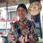 Smiling man in a batik shirt holding three bottled sauces at an outdoor market stall.
