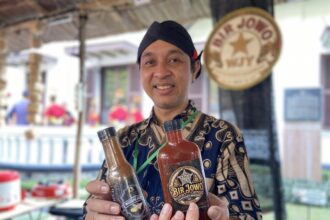 Smiling man in a batik shirt holding three bottled sauces at an outdoor market stall.