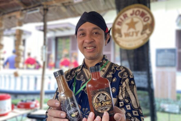 Smiling man in a batik shirt holding three bottled sauces at an outdoor market stall.
