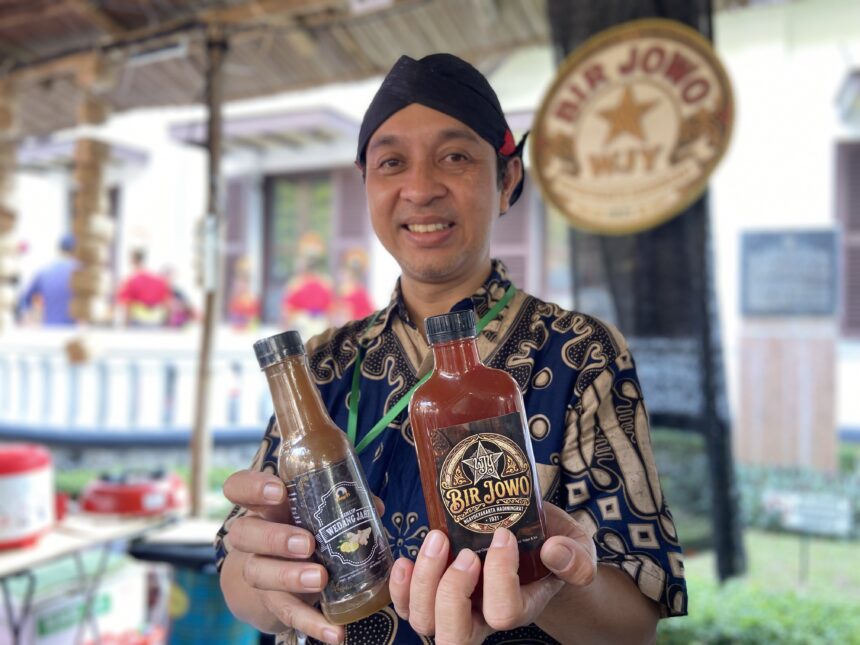 Smiling man in a batik shirt holding three bottled sauces at an outdoor market stall.