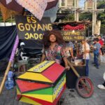 Outdoor street food stall selling es goyang dodol, with colorful stacked boxes and a man with big curly hair in front.