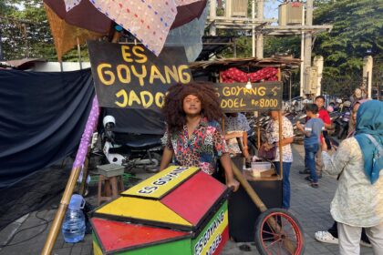 Outdoor street food stall selling es goyang dodol, with colorful stacked boxes and a man with big curly hair in front.
