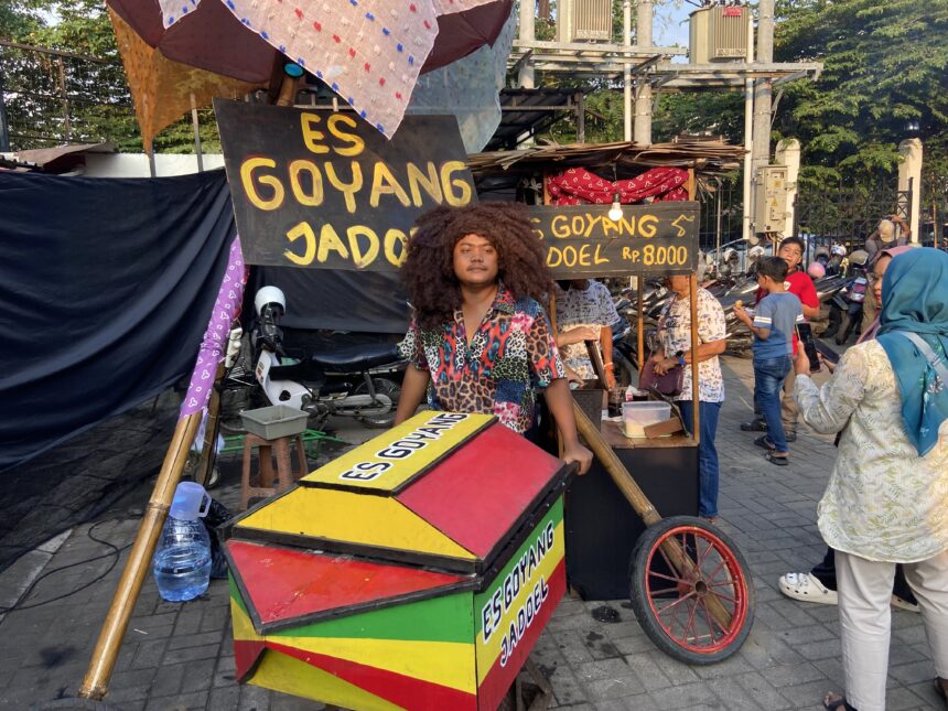 Outdoor street food stall selling es goyang dodol, with colorful stacked boxes and a man with big curly hair in front.