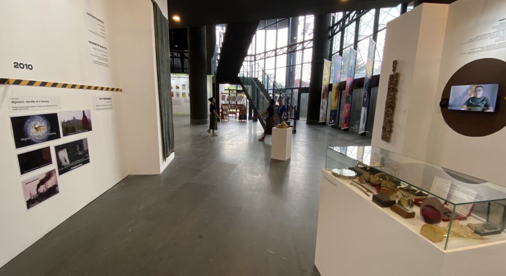 Interior of a modern museum gallery with a 2010 timeline on a white wall to the left, banners along the center, and a glass display case in the foreground right.