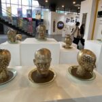 Group of gold-polished bust sculptures with white polka dots arranged on white pedestals in a modern museum gallery, with visitors in the background.”