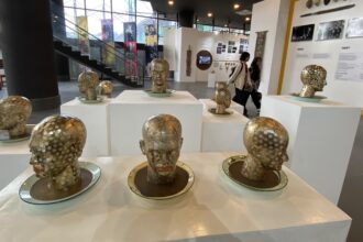 Group of gold-polished bust sculptures with white polka dots arranged on white pedestals in a modern museum gallery, with visitors in the background.”