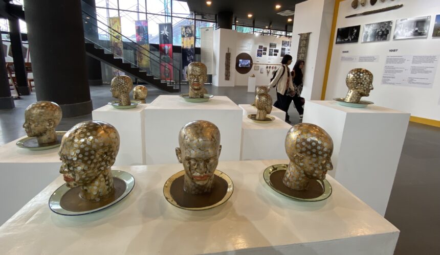 Group of gold-polished bust sculptures with white polka dots arranged on white pedestals in a modern museum gallery, with visitors in the background.”