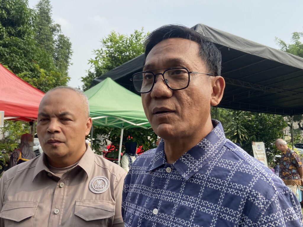 Two men posing for a photo at an outdoor event with colorful tents (red, green) and trees in the background; the man on the right wears glasses and a blue patterned shirt, the man on the left wears a tan uniform.