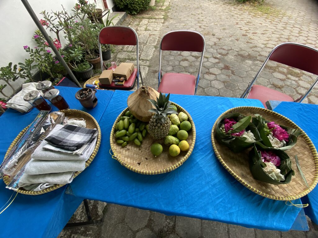 Outdoor market table with blue cloth displaying baskets of fruit (pineapple, limes, mangoes) and flowers, chairs and potted plants in the background.