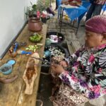 Woman in a bright floral outfit and purple headscarf sits at a wooden table, cutting limes with utensils nearby at an outdoor market setup.