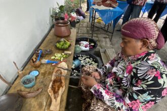 Woman in a bright floral outfit and purple headscarf sits at a wooden table, cutting limes with utensils nearby at an outdoor market setup.