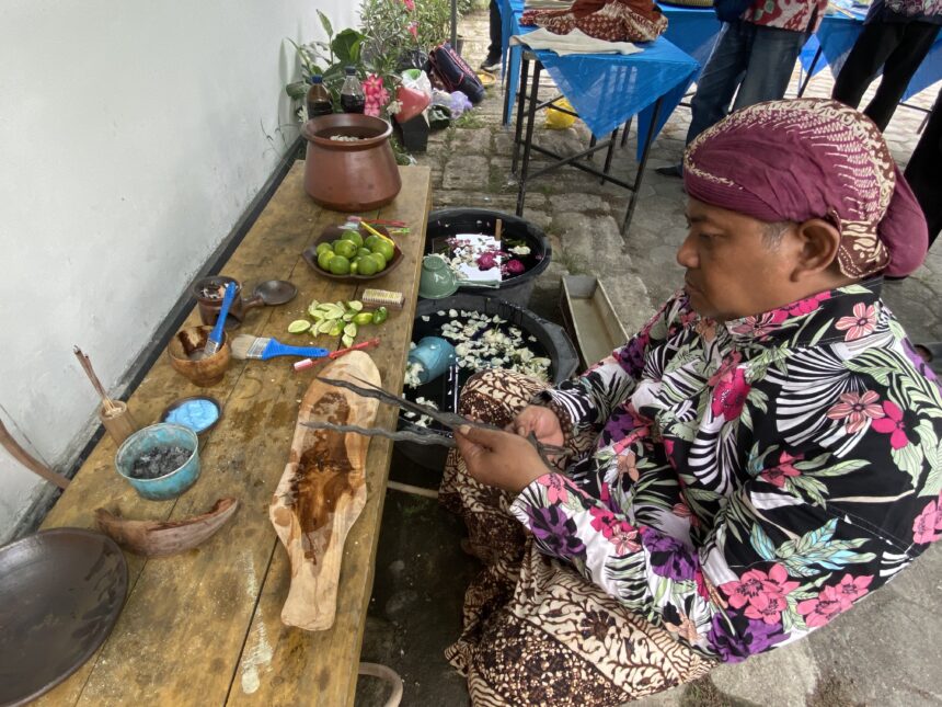 Woman in a bright floral outfit and purple headscarf sits at a wooden table, cutting limes with utensils nearby at an outdoor market setup.