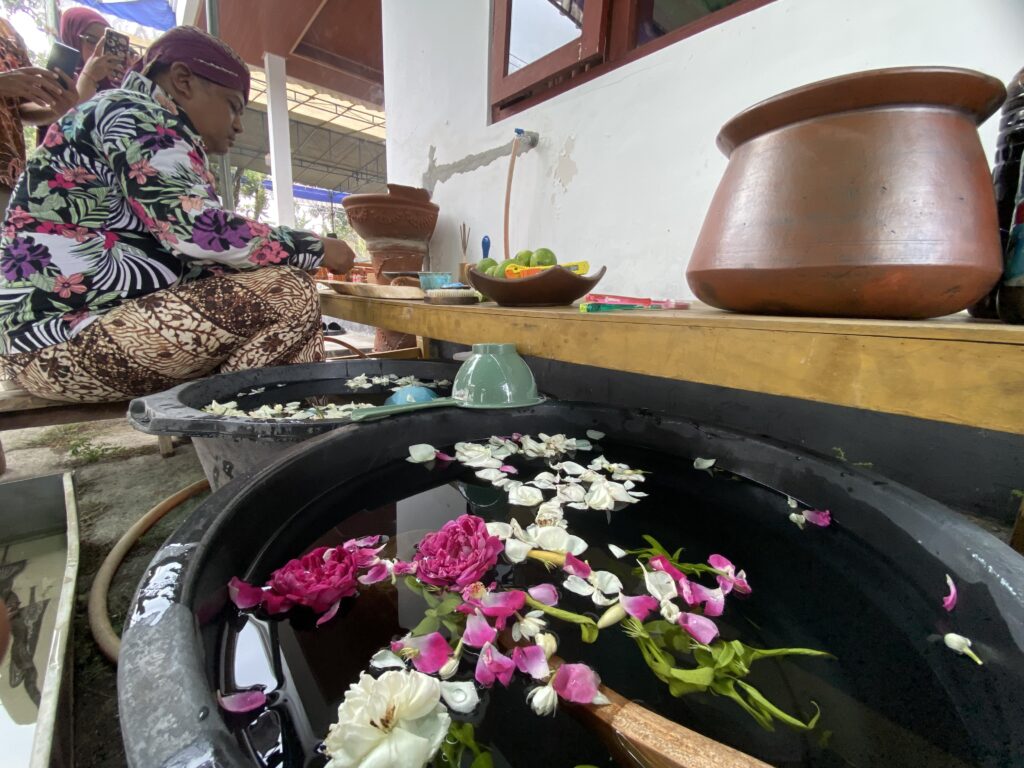 Person in a colorful floral shirt arranges pink and white flower petals in a large water tub, likely making garlands, as onlookers photograph nearby.