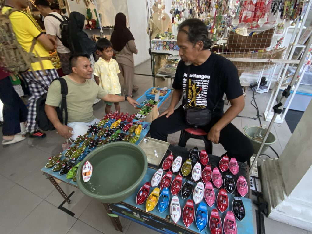 Vendor stall with rows of colorful flip-flops arranged on a table while a seated man points to items and a man stands nearby with a child watching in the background.